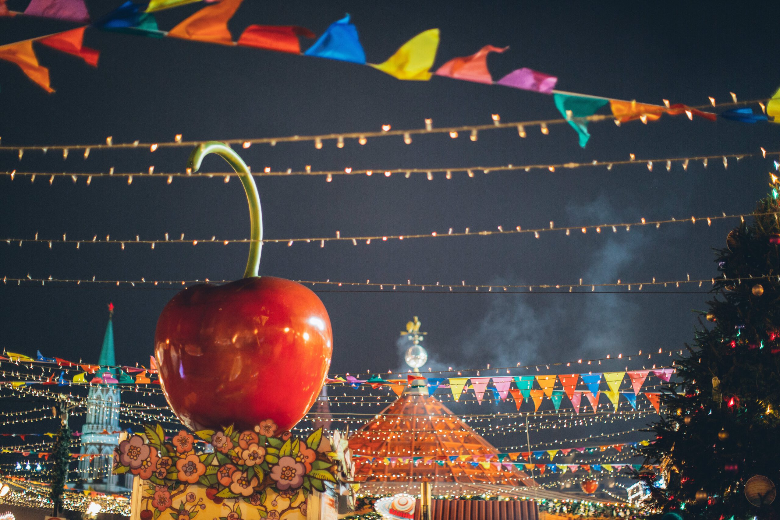 Illustration of Big red glossy toy apple on roof of building on fairground against dark sky in evening city park decorated to winter holidays for the article titled Crash Course in Carnival Celebrations: Tips and Tricks to Host a Fantastic Festivity for Rio Carnival 2024 photographed by Elina Fairytale
