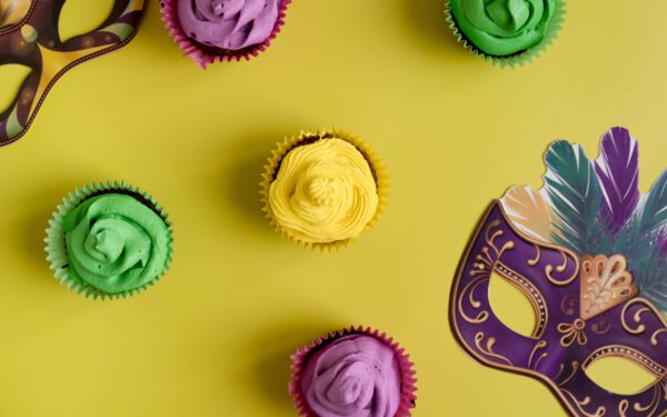 Featured image for the article "The Ultimate Guide to Throwing an Electrifying Mardi Gras Party 2024: Decoration, Costume and Entertainment Ideas". showing Colorful Cupcakes And Masks On Yellow Backg