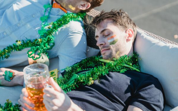 Featured image for the article "Unmasking St. Patrick’s Day 2024: Your Guide to Hosting a Legendary Masquerade Drink Party". showing From above of drunk guy with glass of beer in hands lying on sofa and napping near crop unrecognizable friends after celebrating Feast of Saint Patrick