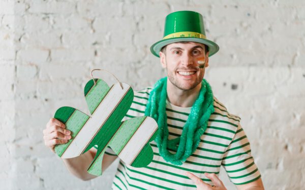Featured image for the article "Preparing for St. Patrick’s Day 2024: Innovative Ideas for Green Decor, Drinks and Party Games". showing Amusing young man with colored in Irish flag cheeks wearing green hat and green sparkle scarf with pasteboard gift in form of Irish symbol shamrock celebrating St Patricks Day