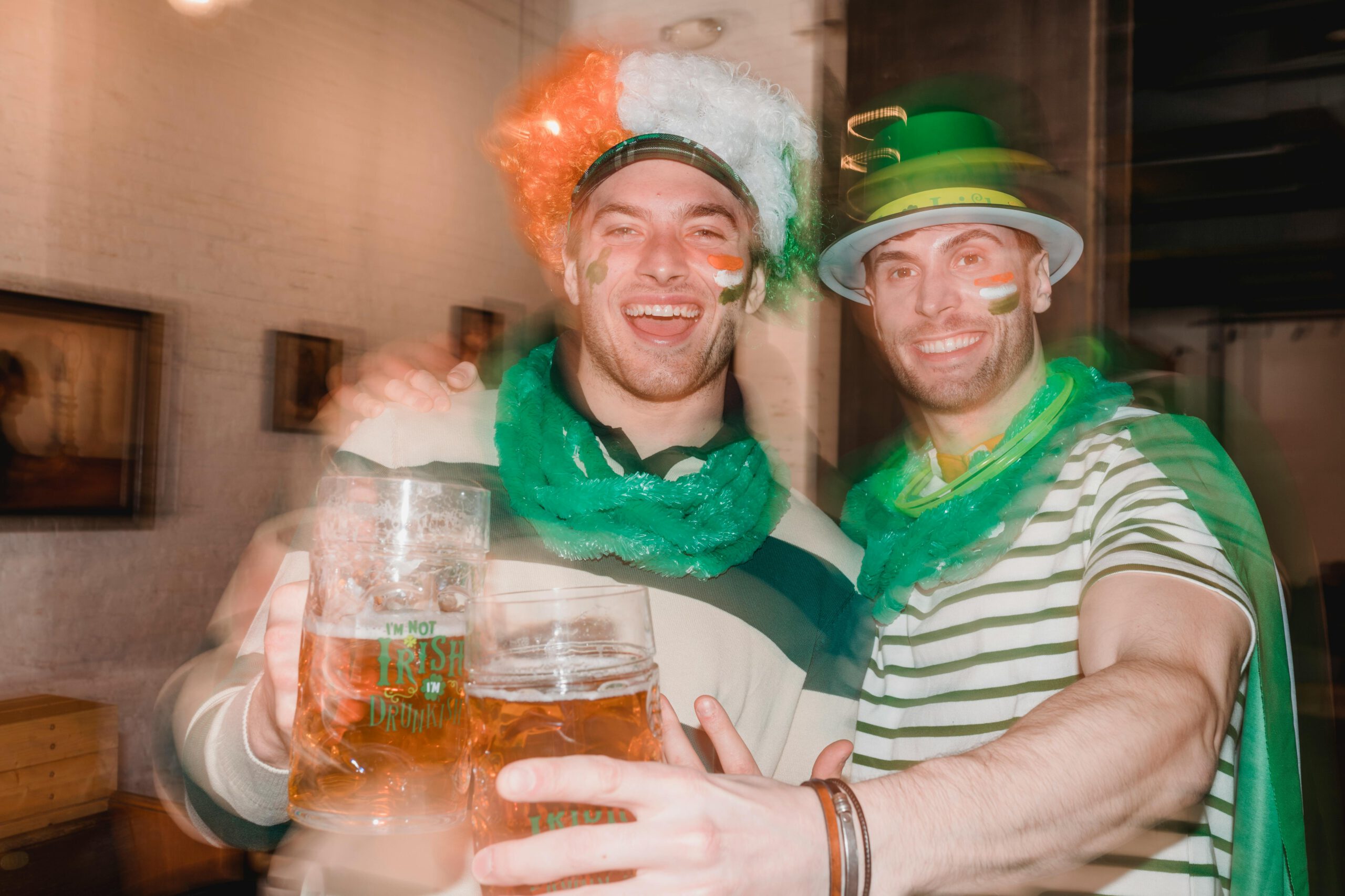 Illustration of Cheerful men covered flag of Ireland and wearing green necklace and green national hat gathering in pub while drinking beer and celebrating Saint Patricks Day for the article titled Unmasking St. Patrick's Day 2024: Your Guide to Hosting a Legendary Masquerade Drink Party photographed by Laura Tancredi