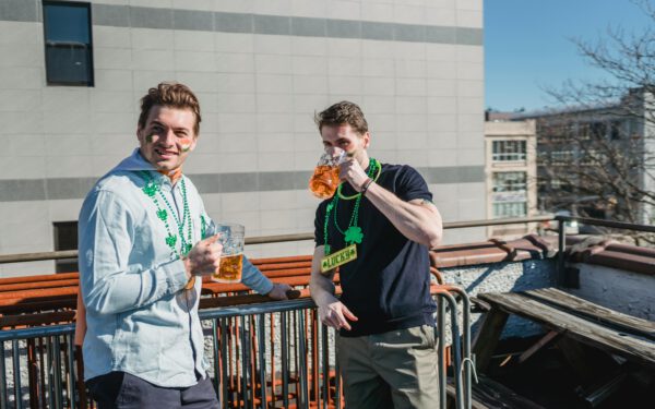Featured image for the article "Spring Parties 2024: The Ultimate Guide to Themed Drink Extravaganzas and Party Games". showing Happy young male millennials in casual clothes and clover necklaces drinking glasses of beer while celebrating Feast of Saint Patrick on sunny day on terrace