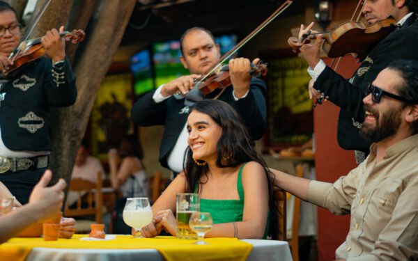 Featured image for the article "Thrill Your Guests with Interactive Cinco de Mayo 2024: An Exciting Guide to Mexican-Themed Drinking Games and Festivities". showing Group of Friends Eating while Listening to Mariachi Band